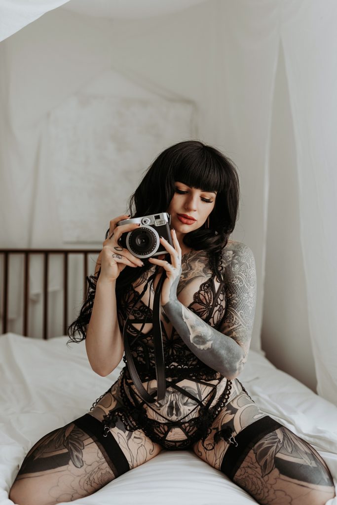 Woman covered in tattoos wearing lingerie kneeling on a bed holding a polaroid camera. 