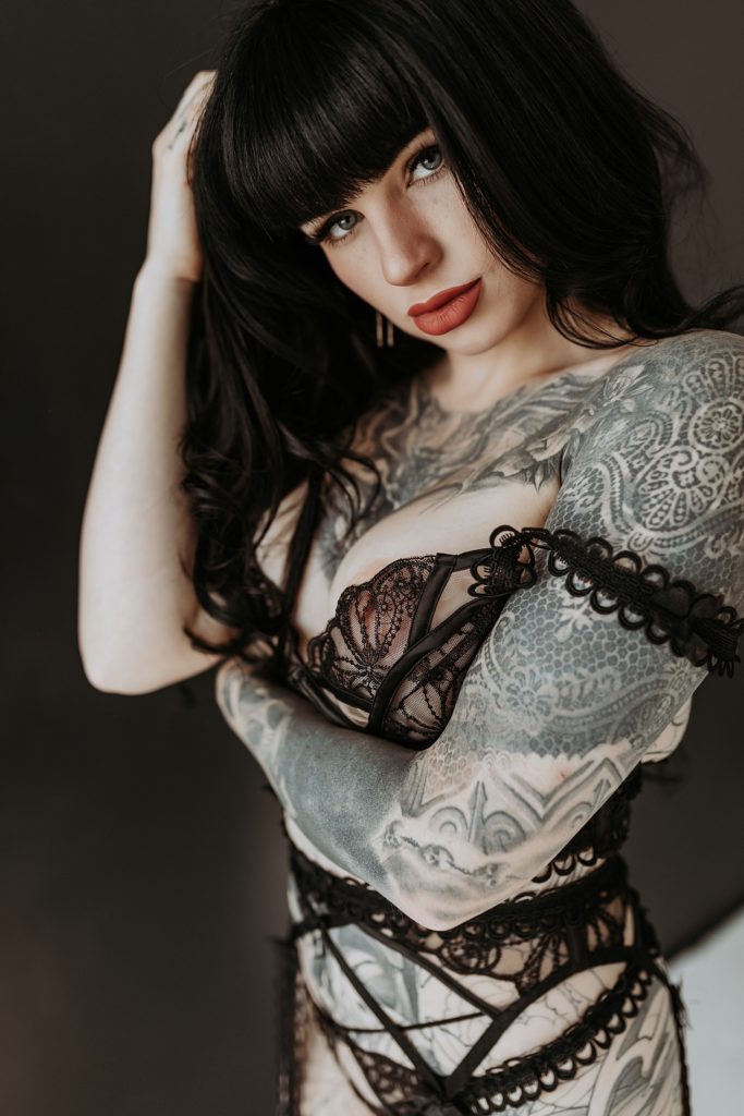 Woman with tattoos in lingerie leaning against a wall, looking at the camera.