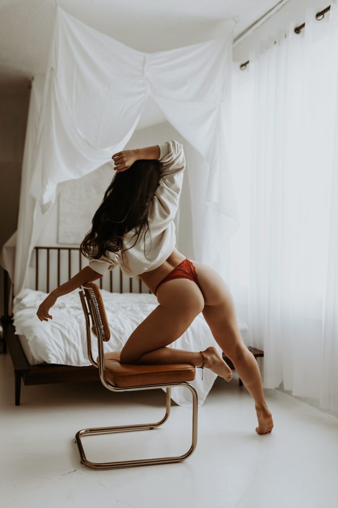 Woman in cream sweater and red lace thong leaning against a chair in the middle of a boudoir studio.