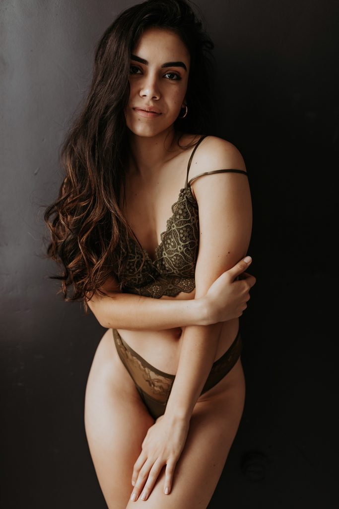 Woman in olive green lingerie leaning against a black wall, looking into the camera.
