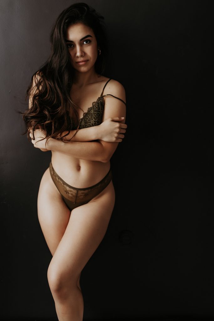 Woman in olive green lingerie leaning against a black wall, looking into the camera.