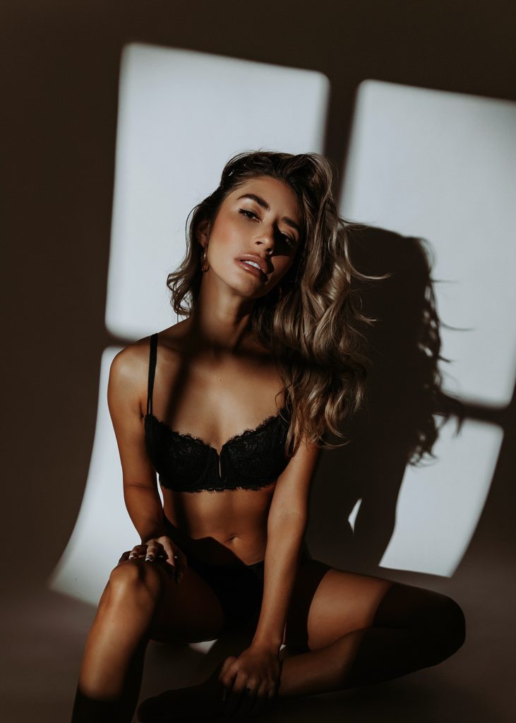 Woman in black lace lingerie in front of a studio backdrop with strobe lights making window shadows fall across her body. 