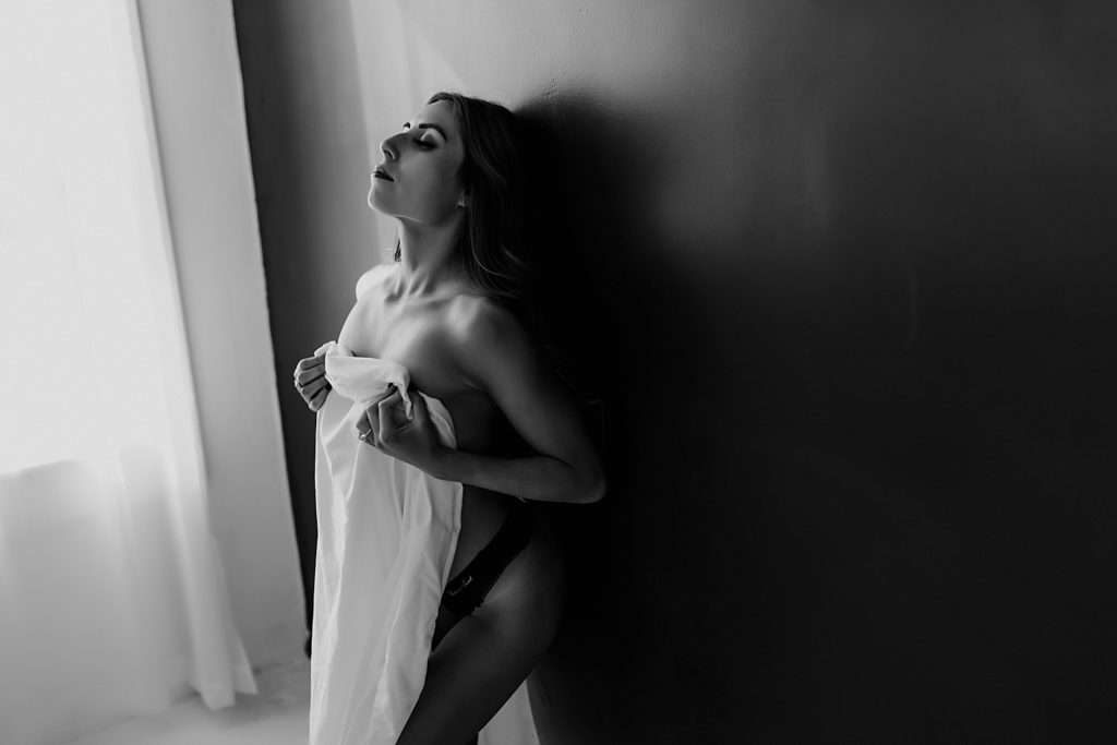 A black and white image of a nude woman leaning against a dark wall in a boudoir studio, covering herself with a sheet. 