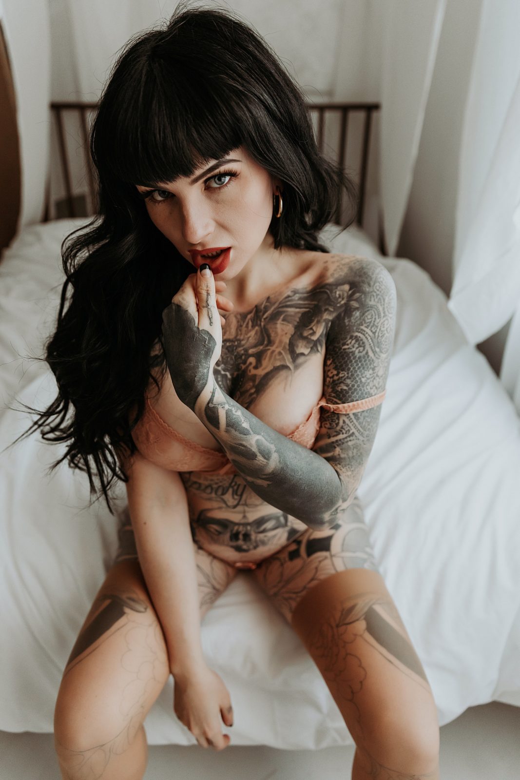 Woman in lingerie sitting on bed with her finger on her lip for her tattoo boudoir session. 
