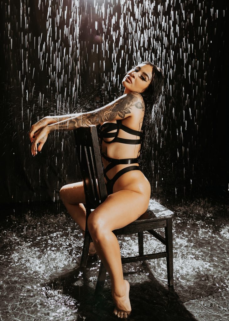 Woman in black lingerie straddling a chair under fake rain for a photo shoot. 