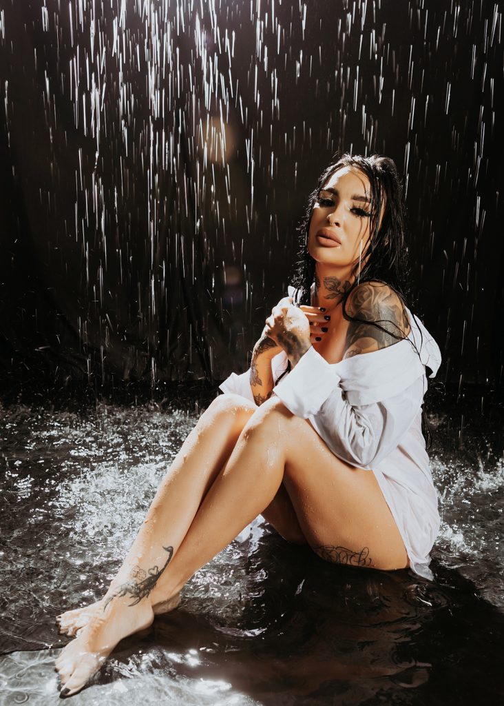 woman with tattoos wearing a white shirt and no pants under a fake rain set. 