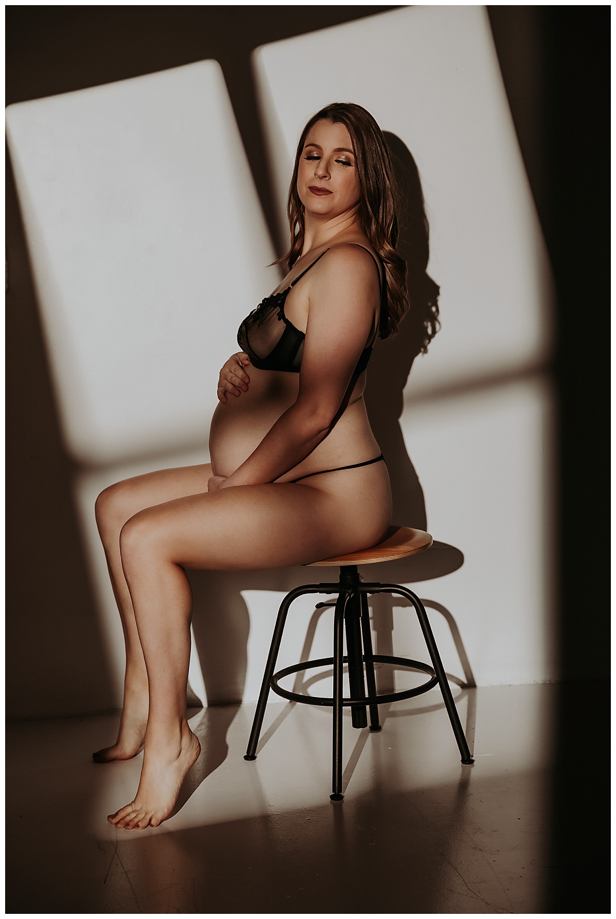 Mama sits on barstool with light shining on pregnant belly for Maternity Boudoir session