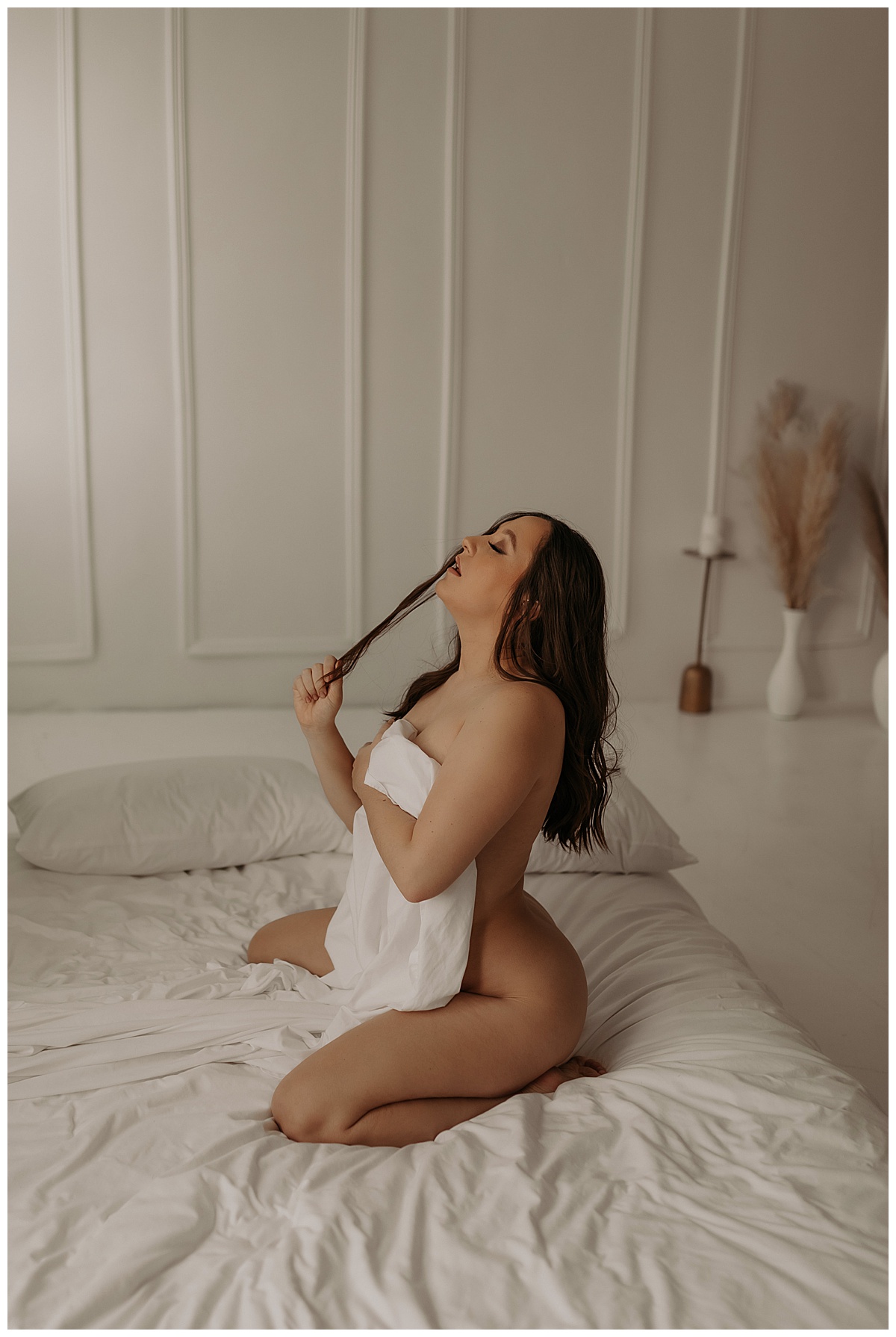 Woman kneels on the bed covering her body with a white sheet showing the benefits of doing More Than One Boudoir Session