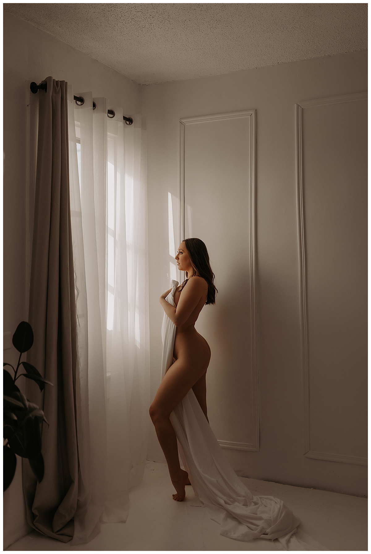 Adult covers her body with a white sheet standing in front of the window for Mary Castillo Photography