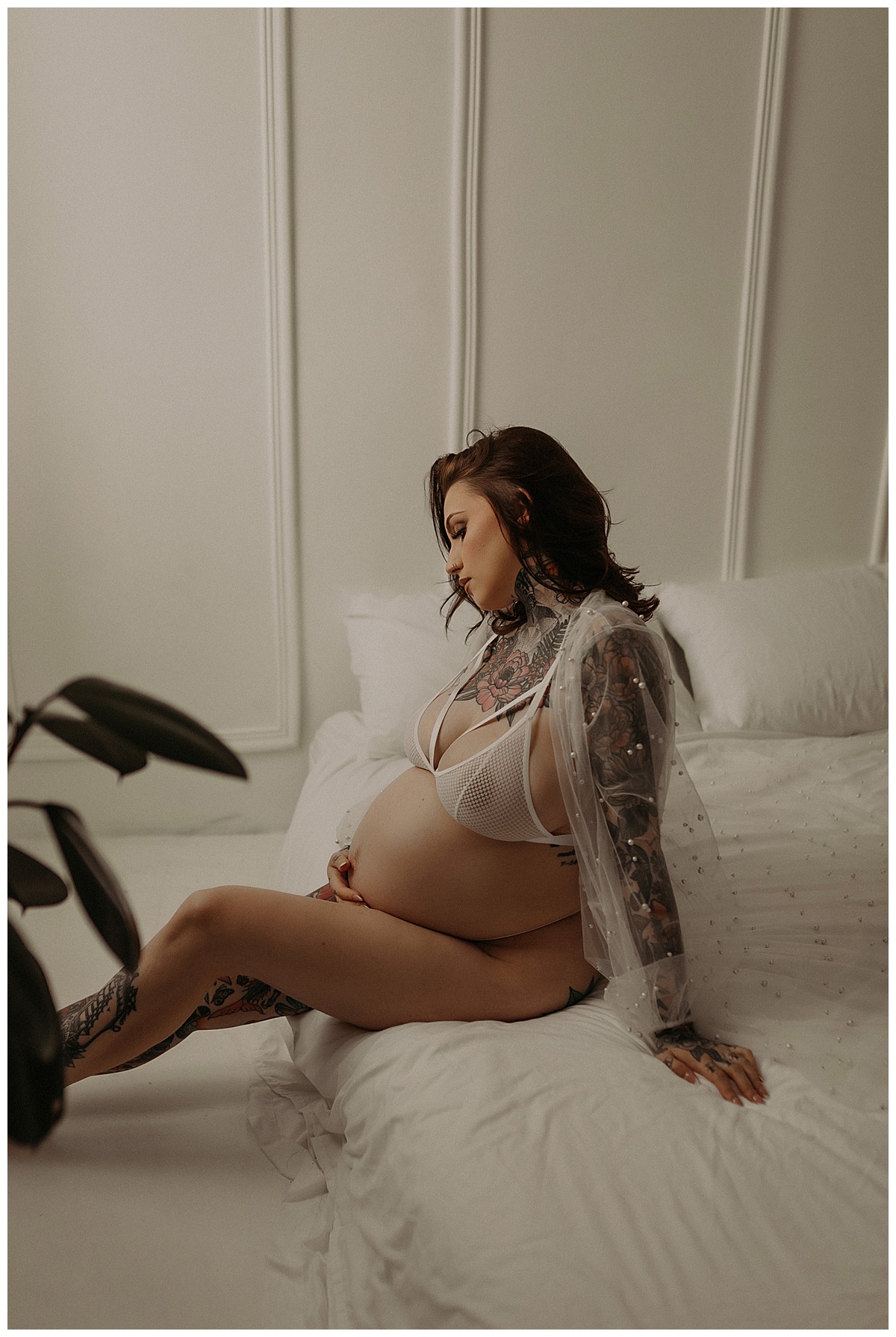 Woman sits on the edge of bed holding her baby bump during her Maternity Boudoir Session