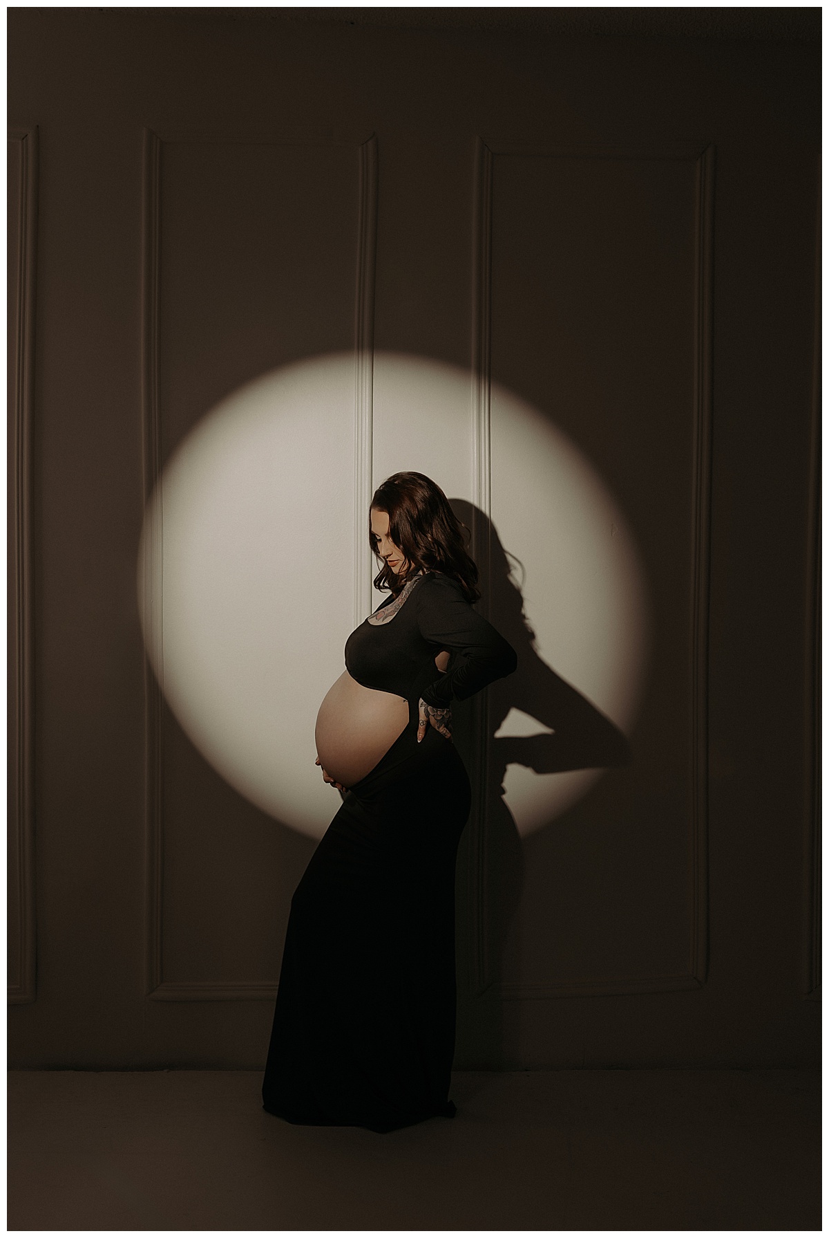 Woman stands in front of spotlight admiring her baby bump during her Maternity Boudoir Session