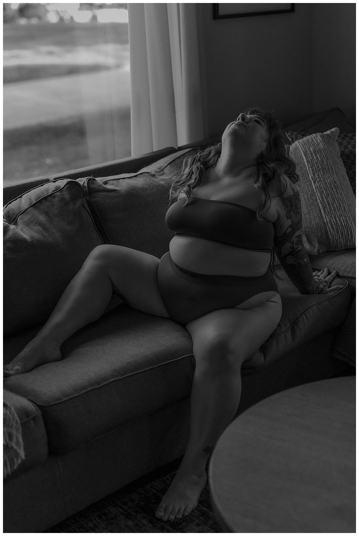 Woman sits on a couch and leans back onto her hands during this Moody Vibes boudoir home session 