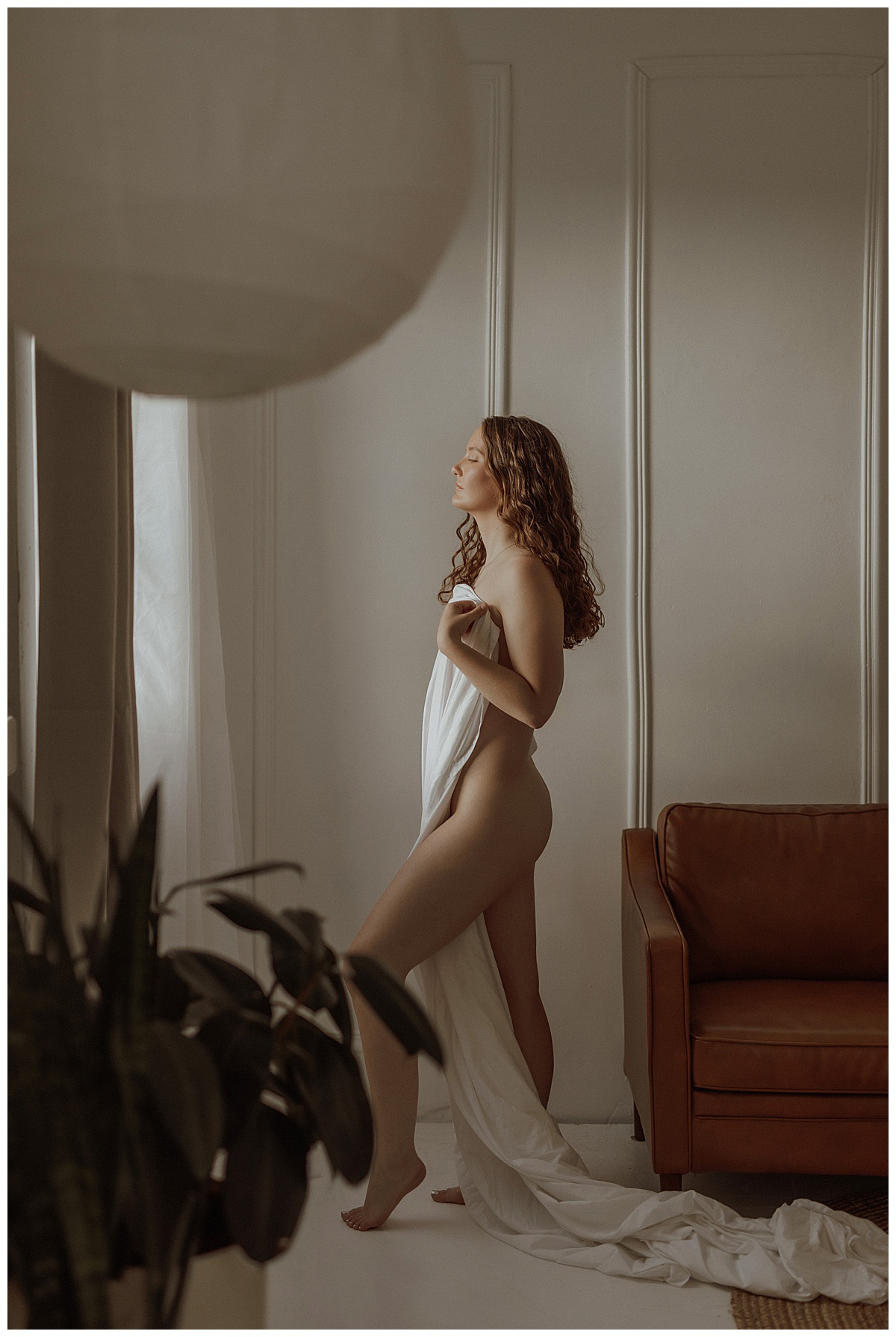 Adult stands in front of the window wrapping a white sheet around her body for Mary Castillo Photography