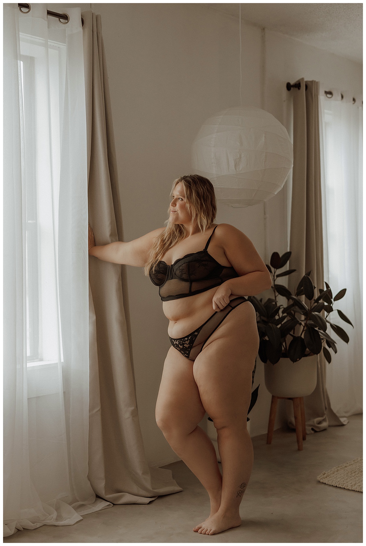 Adult wears black lingerie set standing in front of the window showing why You Deserve This boudoir session