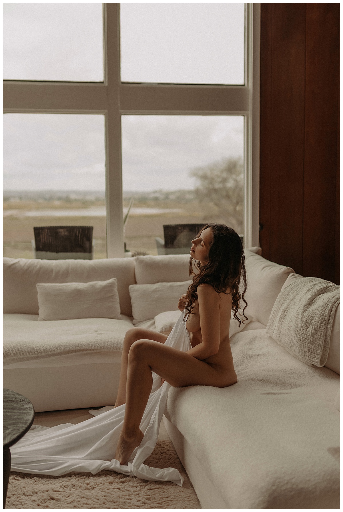 Woman sits down on a couch covering her body with a white sheet for Mary Castillo Photography