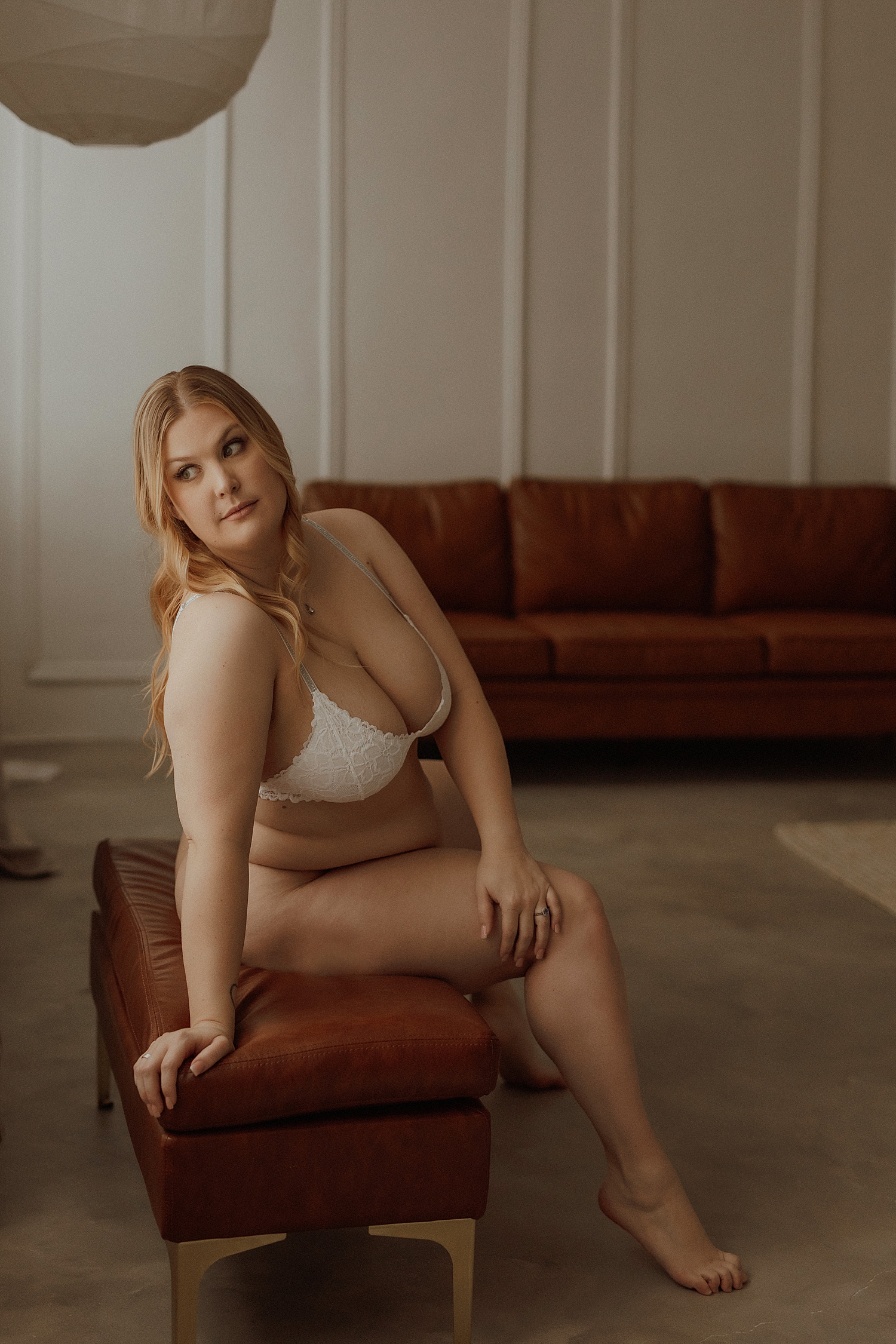 blonde in white sits on leather bench by Minneapolis boudoir photographer 