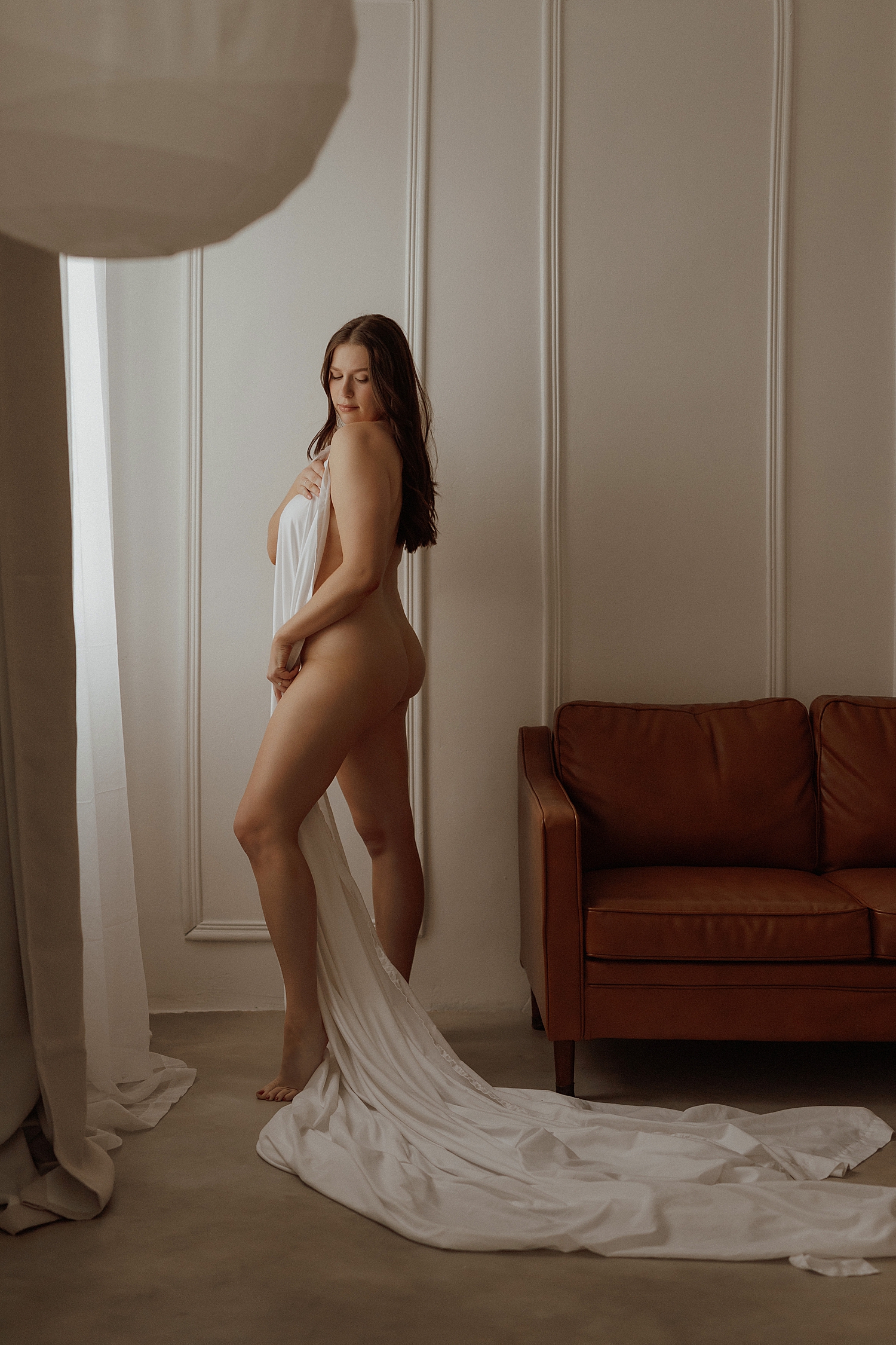brunette stands in front of window holding sheet to cover by Minneapolis photographer