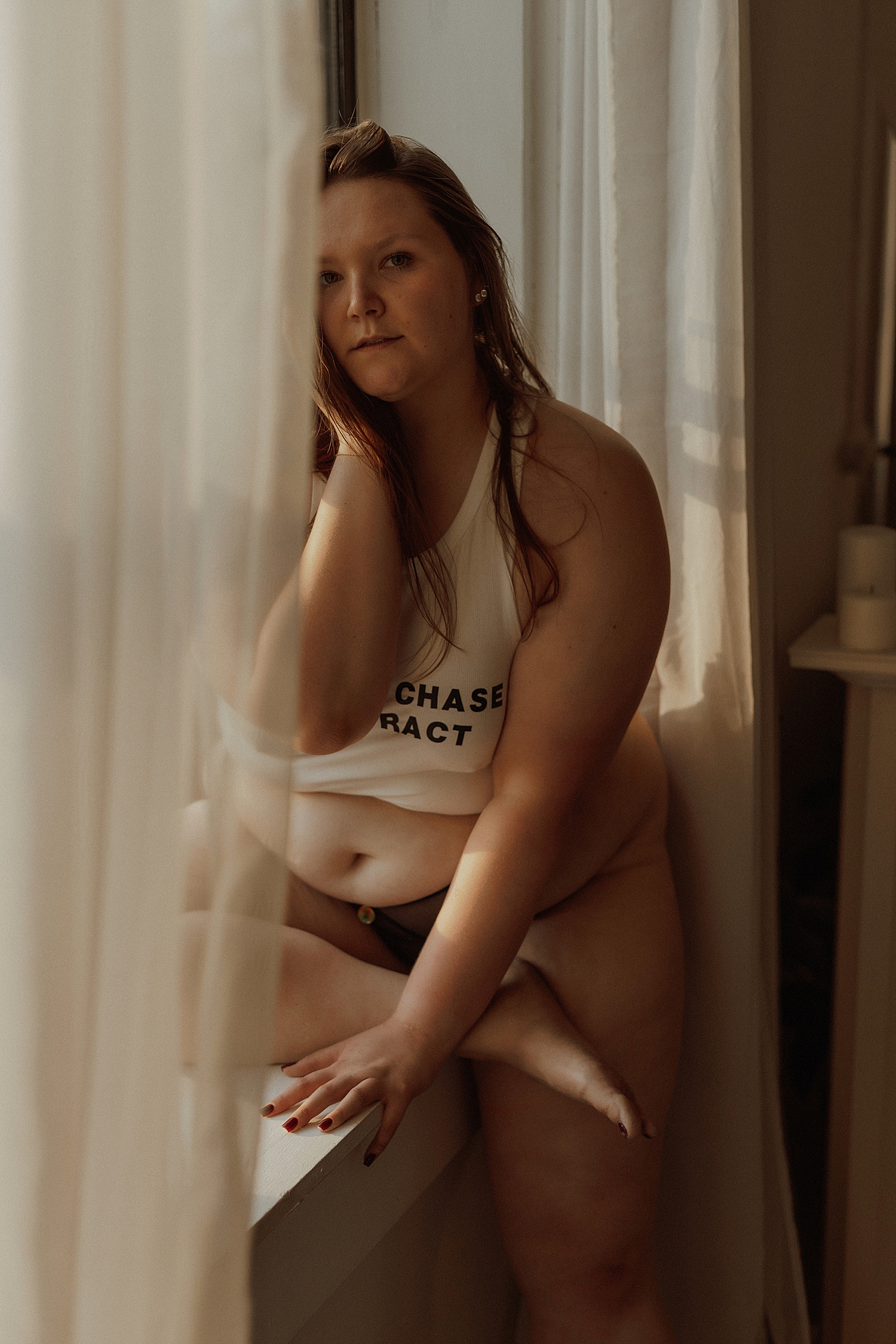 curvy model sits on window pane behind sheer curtains by Minneapolis boudoir photographer