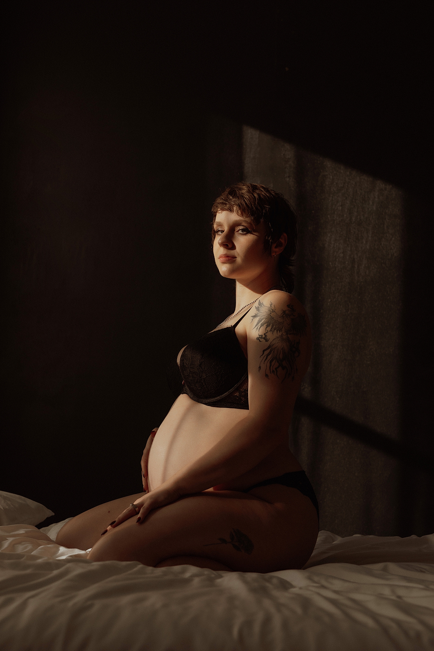 woman in black bra kneels on bed for couples maternity session