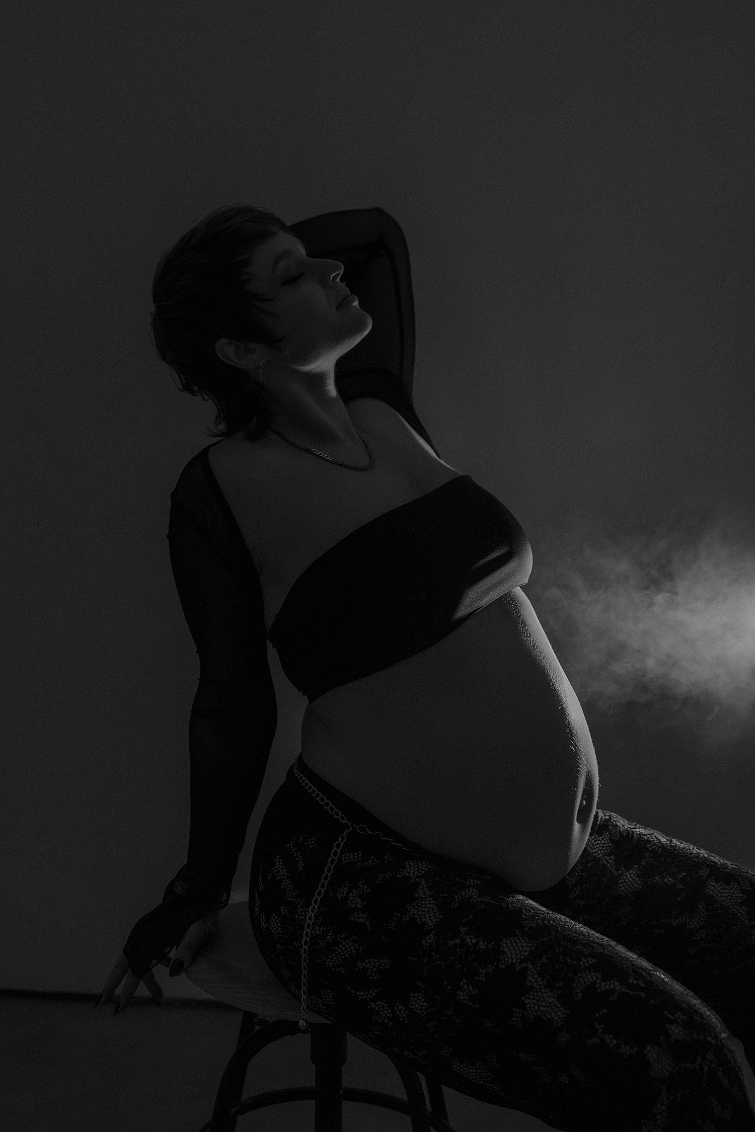 expecting mom leans back on chair with strobe light for couples maternity session